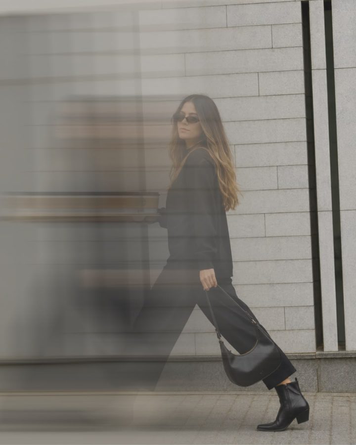 Maria Vitória Tourinho caminhando de óculos escuros e roupa preta, com uma bolsa de couro, diante de uma parede de pedra clara